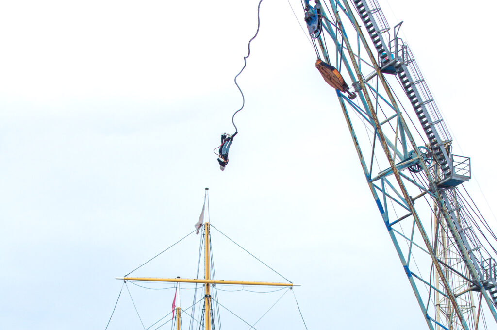 two people doing bungee jumping from crane in front of sailing ship